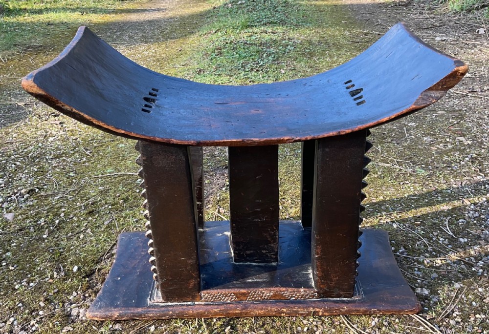A hand-carved dark wood tribal stool featuring a concave seat with pierced slits, central column support, serrated side pillars, and a rectangular base.