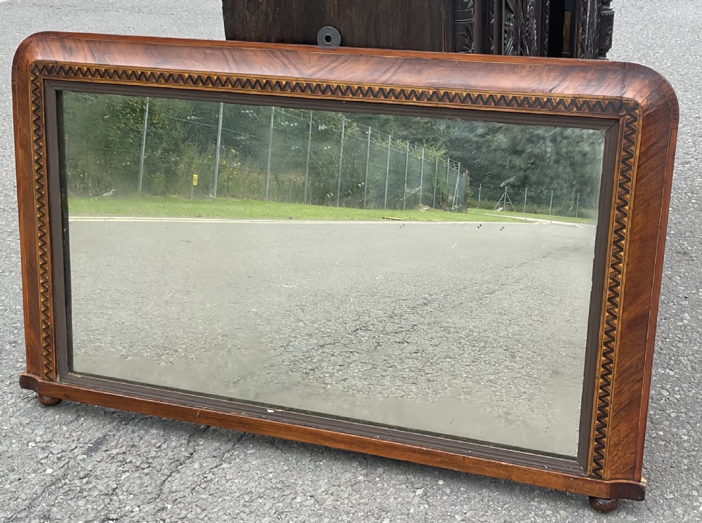 A Victorian rectangular walnut overmantel mirror featuring rounded top corners, geometric marquetry inlay, moulding, and small bun feet.