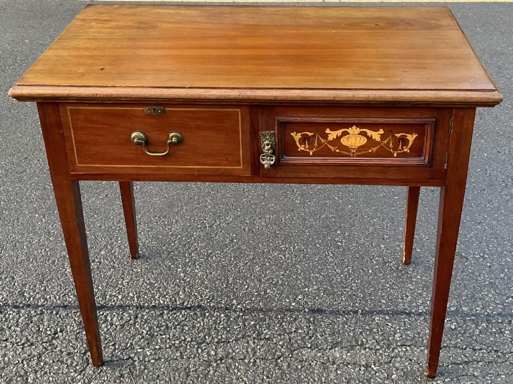 An Edwardian mahogany writing table featuring a flat rectangular top, a single drawer with a brass drop handle, a hinged right panel with floral urn marquetry, and four square tapered legs.