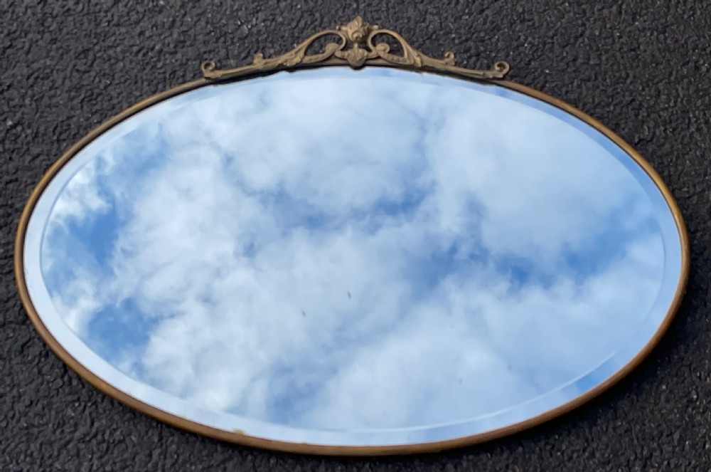 An oval wall mirror featuring a narrow brass frame, a decorative top crest with scrollwork, and a bevelled glass edge.