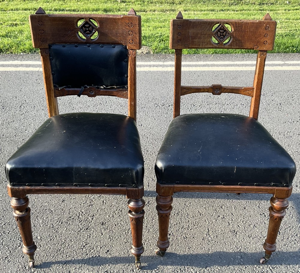 A pair of carved oak Gothic Revival dining chairs featuring black upholstered seats, carved back rails with rosette cut-outs, turned front legs, and brass castors.