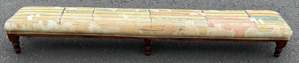 An elongated Edwardian upholstered footstool featuring six turned timber legs and patterned yellow fabric upholstery.