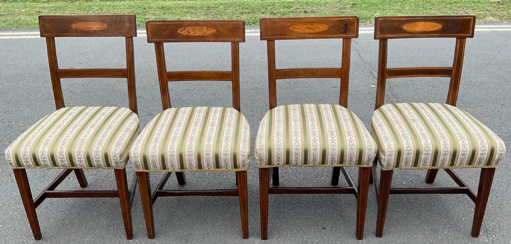 A set of four antique Georgian mahogany dining chairs featuring oval satinwood crest rail inlays, square tapered legs with stretchers, and striped green and cream fabric seats.