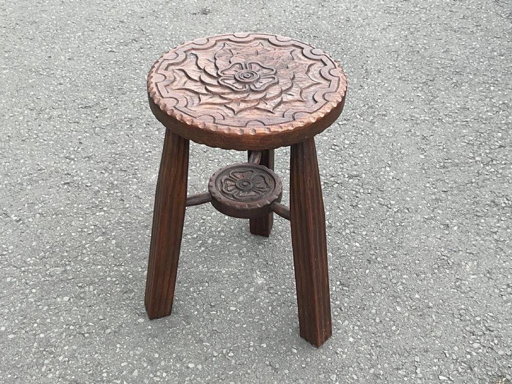 A dark carved oak three-legged circular stool featuring a central floral motif carved on the top seat and a lower stretcher assembly.