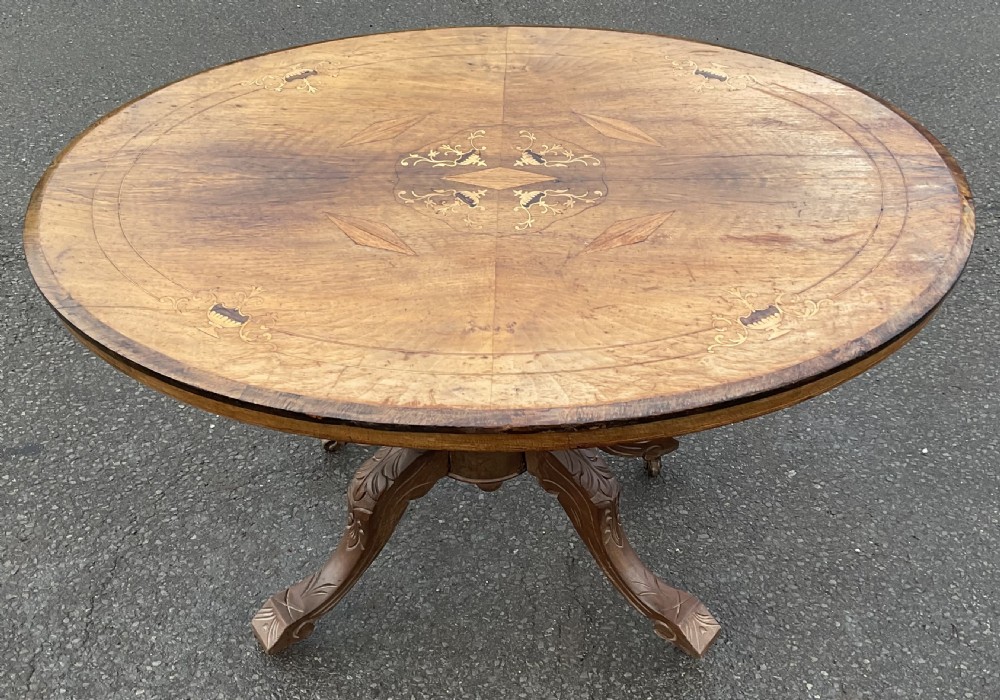 An oval walnut centre table featuring floral marquetry inlays on the top surface and a carved four-legged pedestal base.