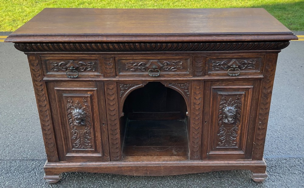 Victorian dark oak sideboard featuring three carved drawers, two carved cupboard doors with relief face motifs, and a central open arch.