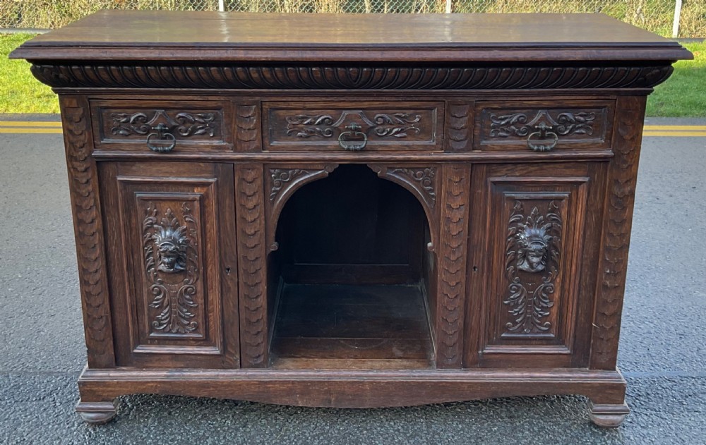 Victorian dark oak sideboard featuring three carved drawers, two carved cupboard doors with relief face motifs, and a central open arch. View III