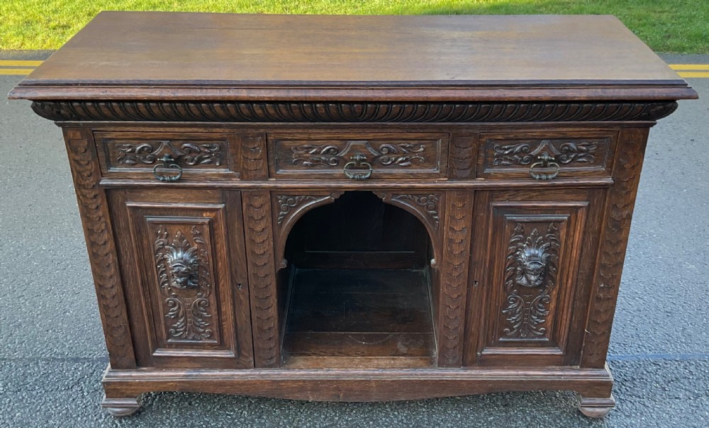 Victorian dark oak sideboard featuring three carved drawers, two carved cupboard doors with relief face motifs, and a central open arch. View VI