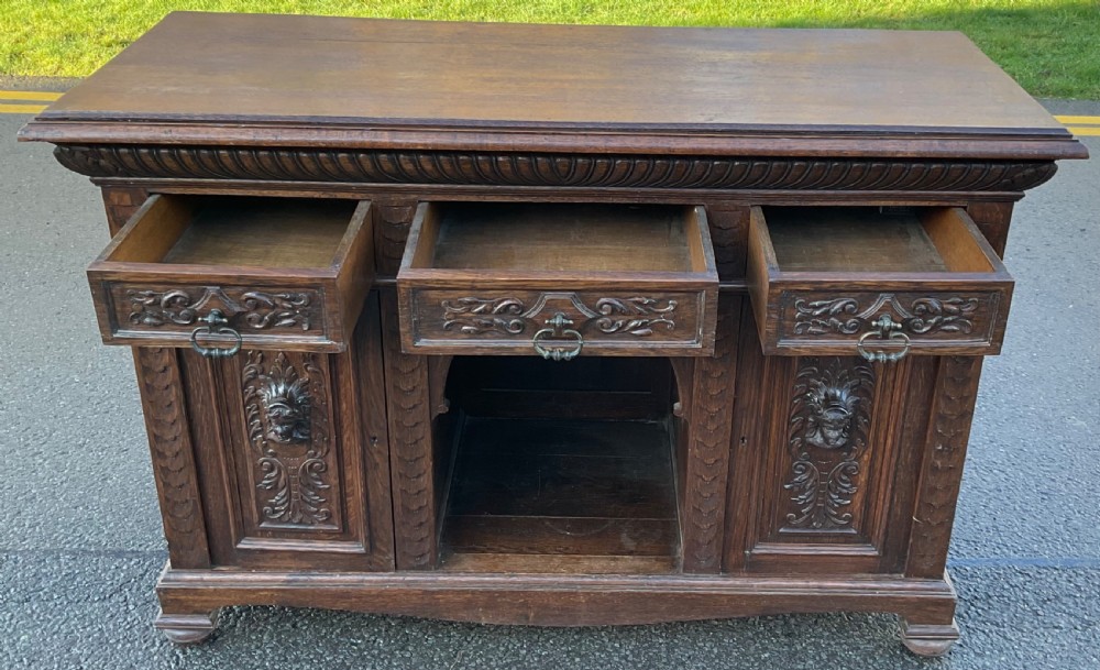 Victorian dark oak sideboard featuring three carved drawers, two carved cupboard doors with relief face motifs, and a central open arch. View VII