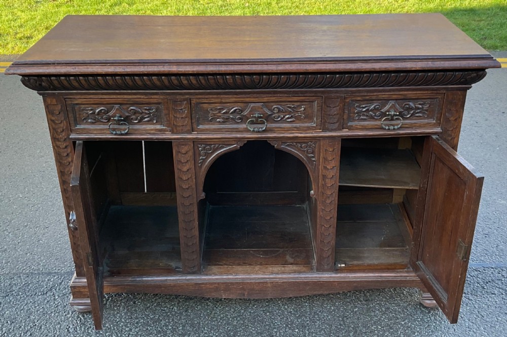 Victorian dark oak sideboard featuring three carved drawers, two carved cupboard doors with relief face motifs, and a central open arch. View VIII