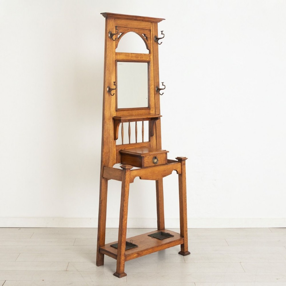 An Arts and Crafts golden oak hall stand featuring a central mirror, top coat hooks, a small glove drawer, and lower umbrella drip trays.