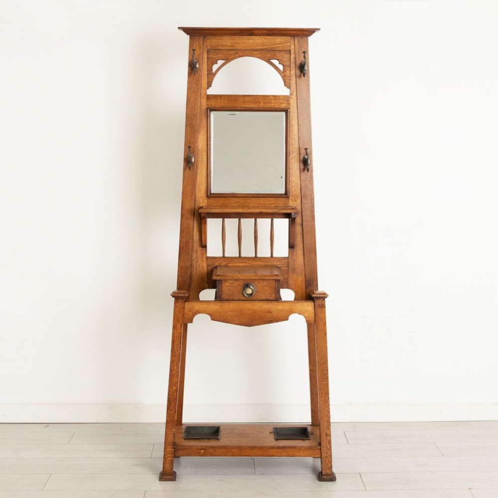 An Arts and Crafts golden oak hall stand featuring a central mirror, top coat hooks, a small glove drawer, and lower umbrella drip trays. View II