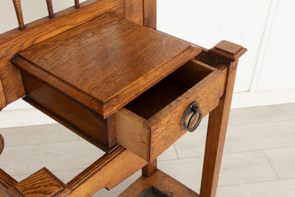 An Arts and Crafts golden oak hall stand featuring a central mirror, top coat hooks, a small glove drawer, and lower umbrella drip trays. View III