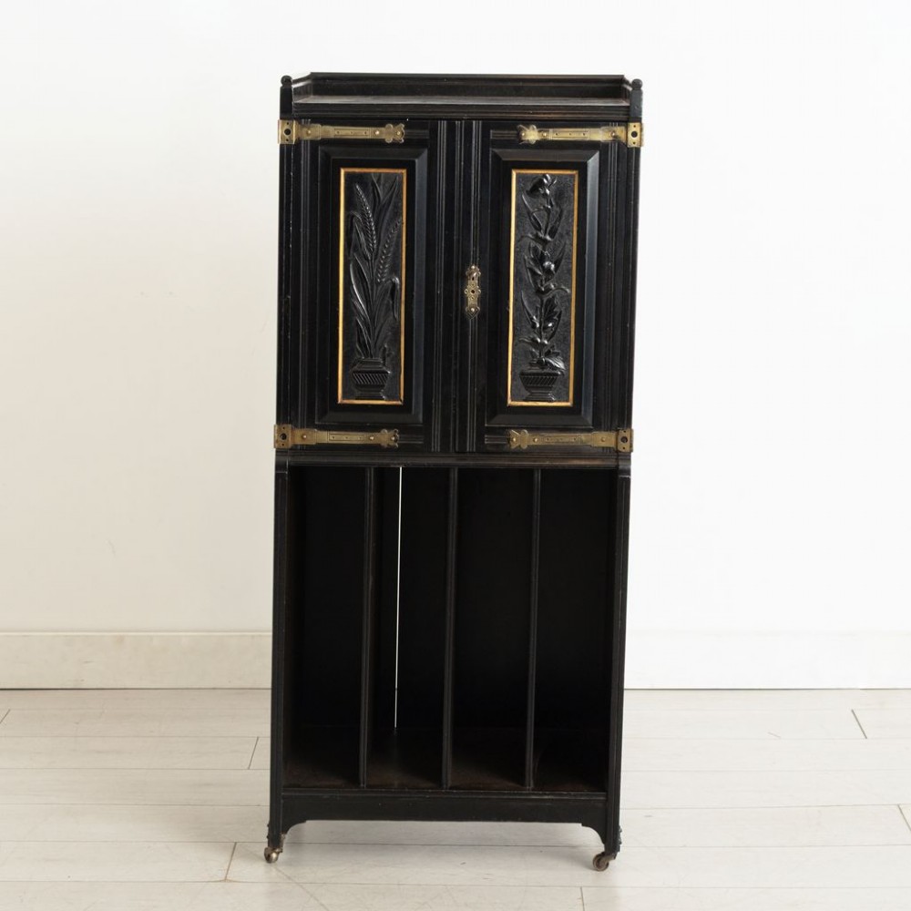 A late 19th-century Aesthetic Movement ebonised music cabinet featuring carved botanical upper doors with brass corner mounts and open vertical sheet music slots below. View II