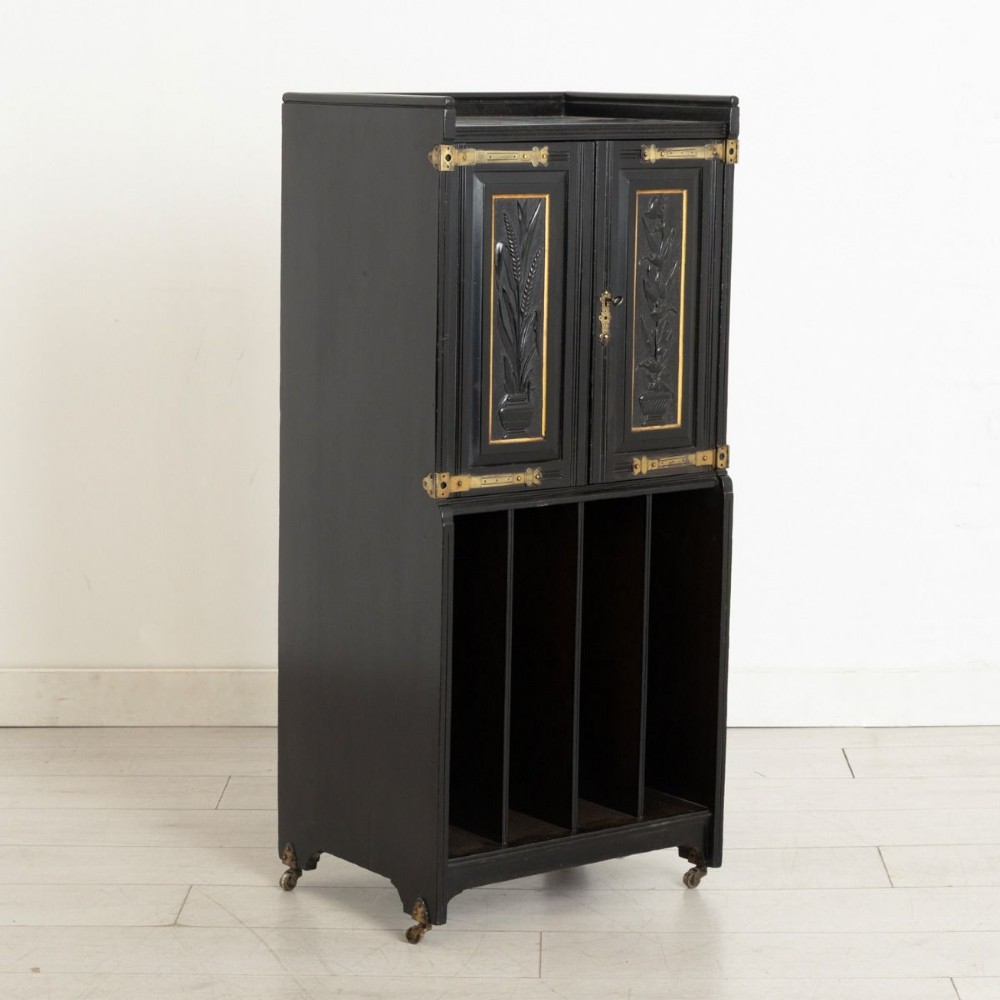 A late 19th-century Aesthetic Movement ebonised music cabinet featuring carved botanical upper doors with brass corner mounts and open vertical sheet music slots below.