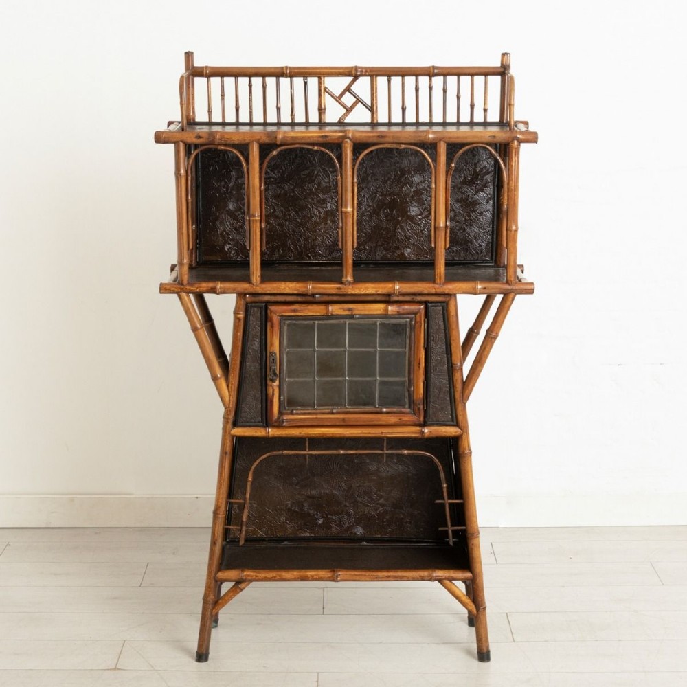 An antique Victorian bamboo display cabinet featuring an upper gallery shelf, open compartments with dark embossed panels, and a central leaded glass door.