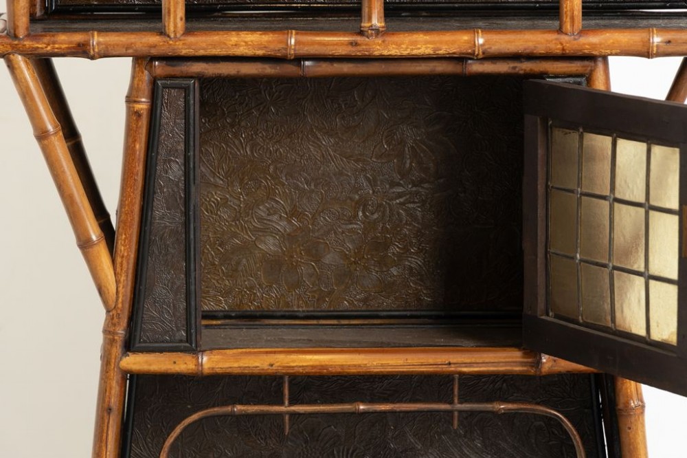 An antique Victorian bamboo display cabinet featuring an upper gallery shelf, open compartments with dark embossed panels, and a central leaded glass door. View IV