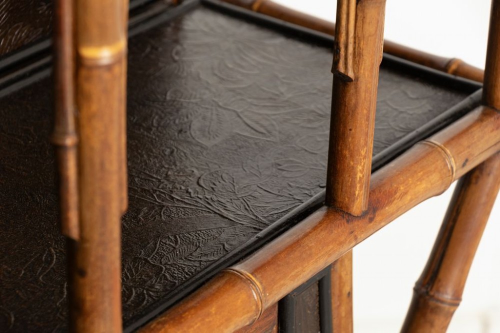 An antique Victorian bamboo display cabinet featuring an upper gallery shelf, open compartments with dark embossed panels, and a central leaded glass door. View V