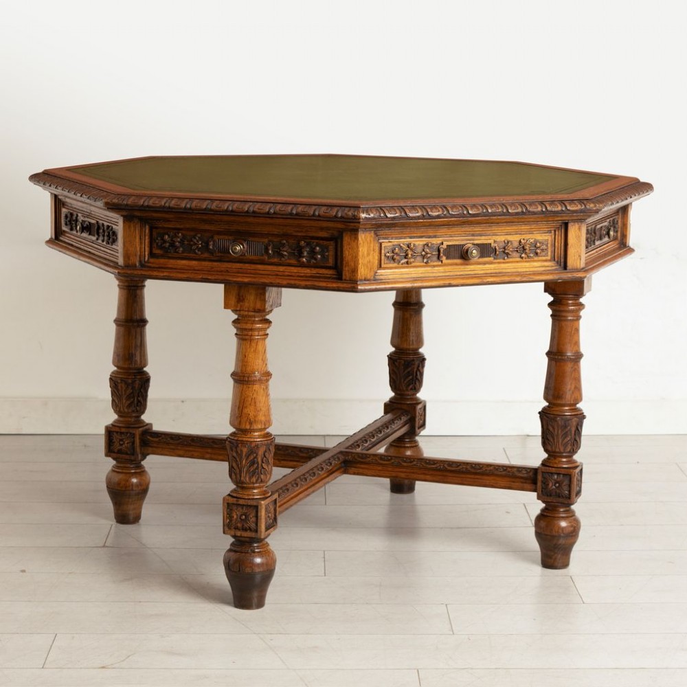 An antique Victorian octagonal oak library table with a green leather inset writing top, relief-carved frieze drawers, turned legs, and a carved X-stretcher.