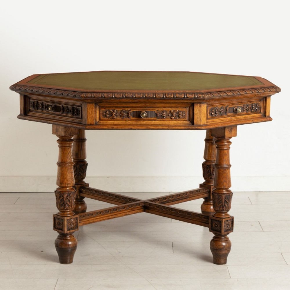 An antique Victorian octagonal oak library table with a green leather inset writing top, relief-carved frieze drawers, turned legs, and a carved X-stretcher. View II