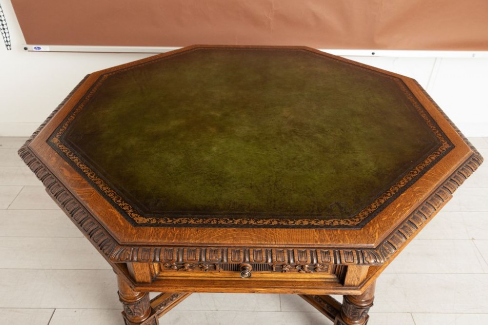 An antique Victorian octagonal oak library table with a green leather inset writing top, relief-carved frieze drawers, turned legs, and a carved X-stretcher. View V