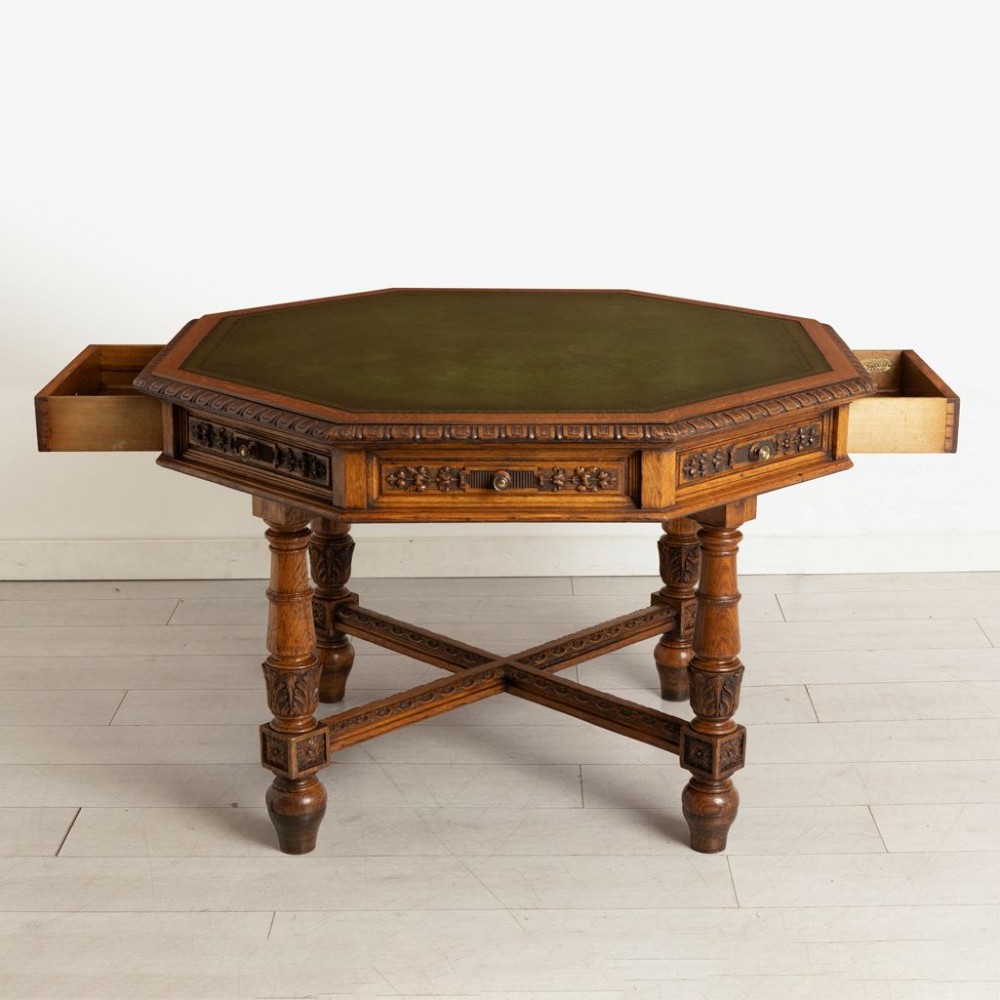 An antique Victorian octagonal oak library table with a green leather inset writing top, relief-carved frieze drawers, turned legs, and a carved X-stretcher. View VII