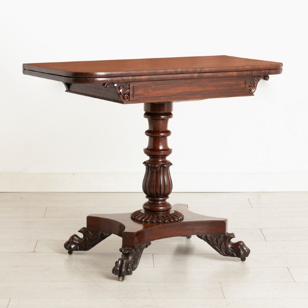 A Late Georgian mahogany fold-over card table featuring a turned carved pedestal stem and a quadform base with carved scroll feet on castors.