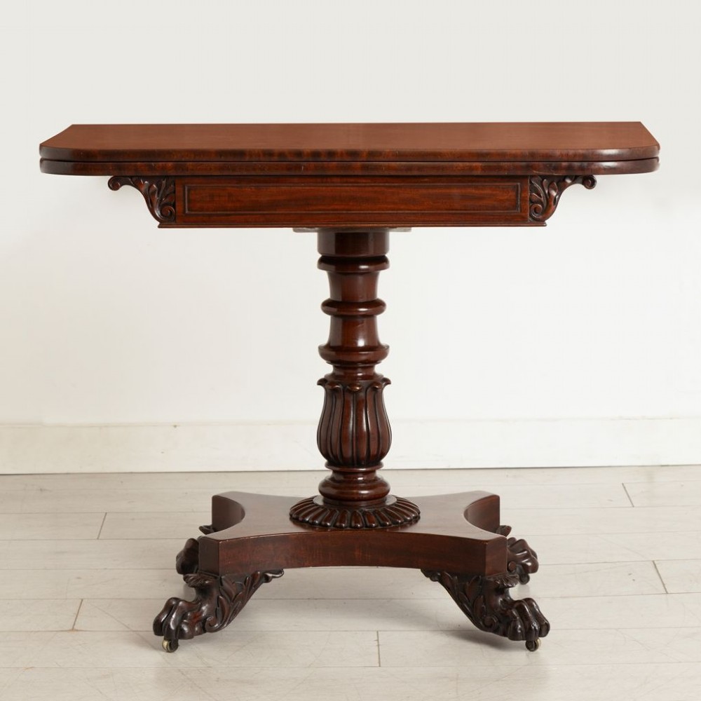 A Late Georgian mahogany fold-over card table featuring a turned carved pedestal stem and a quadform base with carved scroll feet on castors. View II