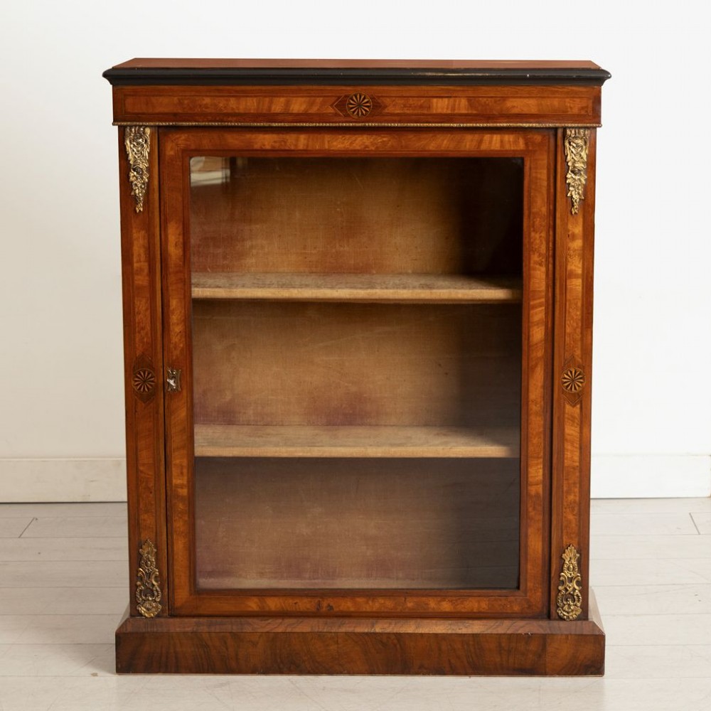 A Victorian burr walnut pier cabinet featuring a single glazed door, ebony and rosewood inlay detailing, interior wooden shelves, and gilt brass ormolu corner mounts.