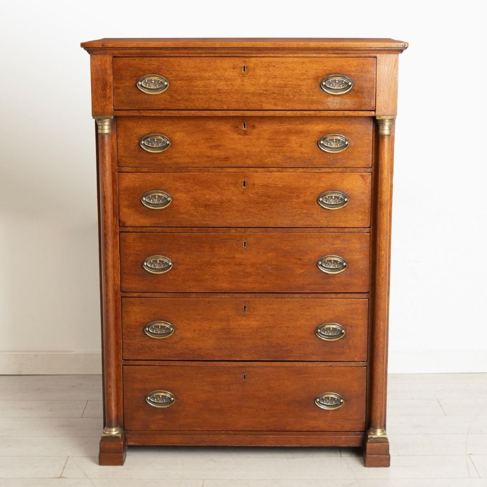 A tall mid-19th century French oak chest of drawers featuring six stacked drawers, brass oval drop handles, keyhole escutcheons, and flanking columns with brass accents.