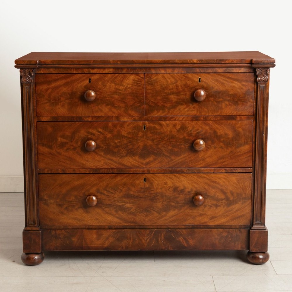 An antique mahogany chest of drawers featuring flame mahogany veneer fronts, turned knobs, side pilasters with leaf carvings, and bun feet.