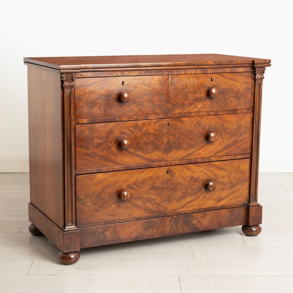 An antique mahogany chest of drawers featuring flame mahogany veneer fronts, turned knobs, side pilasters with leaf carvings, and bun feet. View II