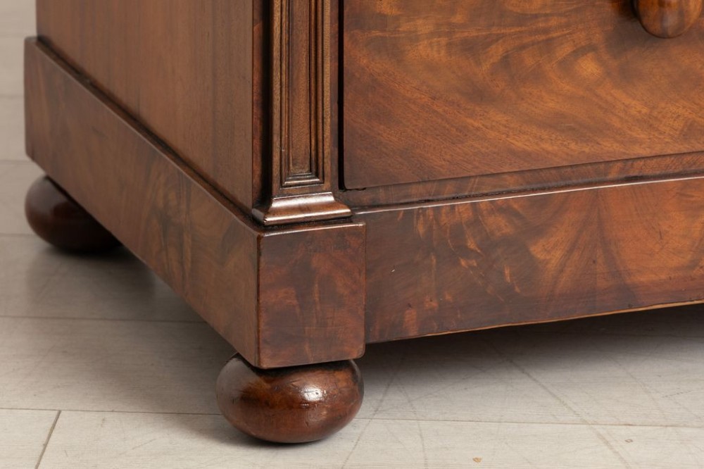 An antique mahogany chest of drawers featuring flame mahogany veneer fronts, turned knobs, side pilasters with leaf carvings, and bun feet. View III
