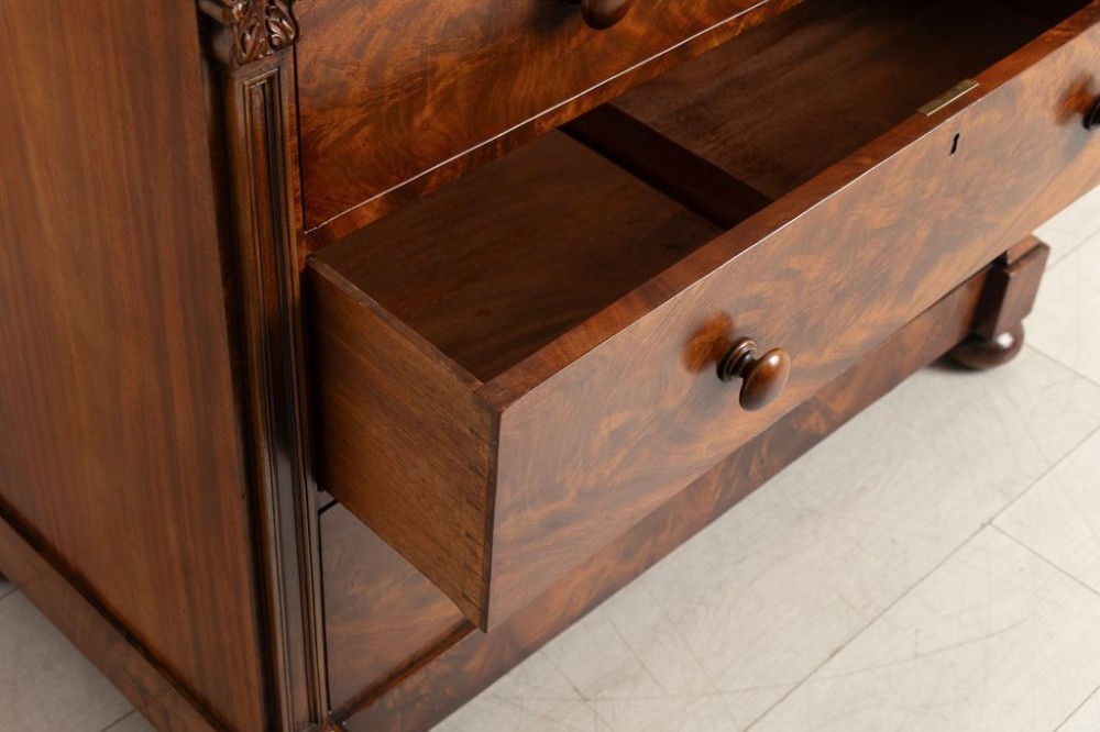 An antique mahogany chest of drawers featuring flame mahogany veneer fronts, turned knobs, side pilasters with leaf carvings, and bun feet. View IV