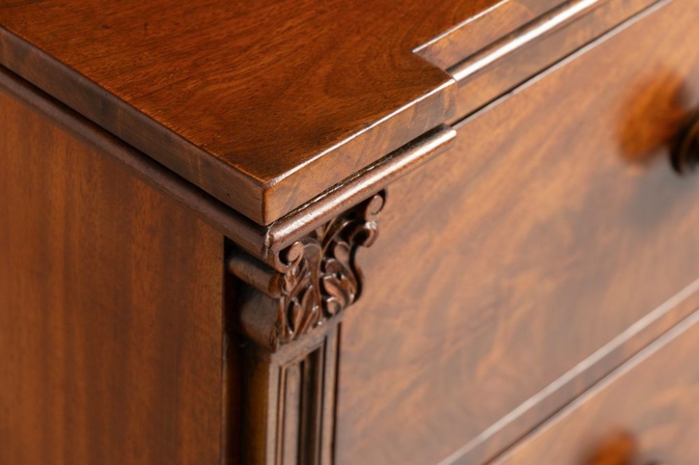 An antique mahogany chest of drawers featuring flame mahogany veneer fronts, turned knobs, side pilasters with leaf carvings, and bun feet. View V
