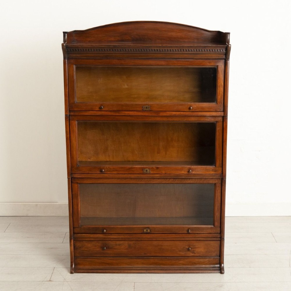 An Edwardian oak three-tier sectional bookcase featuring up-and-over glazed doors, an arched pediment top, and a base drawer with wooden knobs.