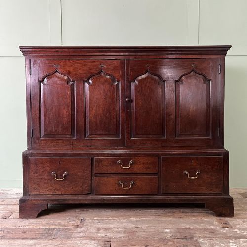 fig 1. An 18th-century dark oak low linen press cupboard featuring arched fielded panel doors over a base with four drawers and brass handles.
