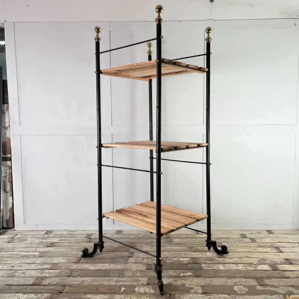A tall Victorian-style black metal frame display stand featuring three timber slatted shelves, decorative brass finials at the top corners, and scrolled foot supports.