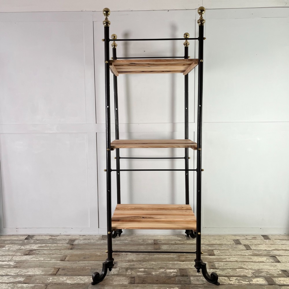 A tall Victorian-style black metal frame display stand featuring three timber slatted shelves, decorative brass finials at the top corners, and scrolled foot supports. View II