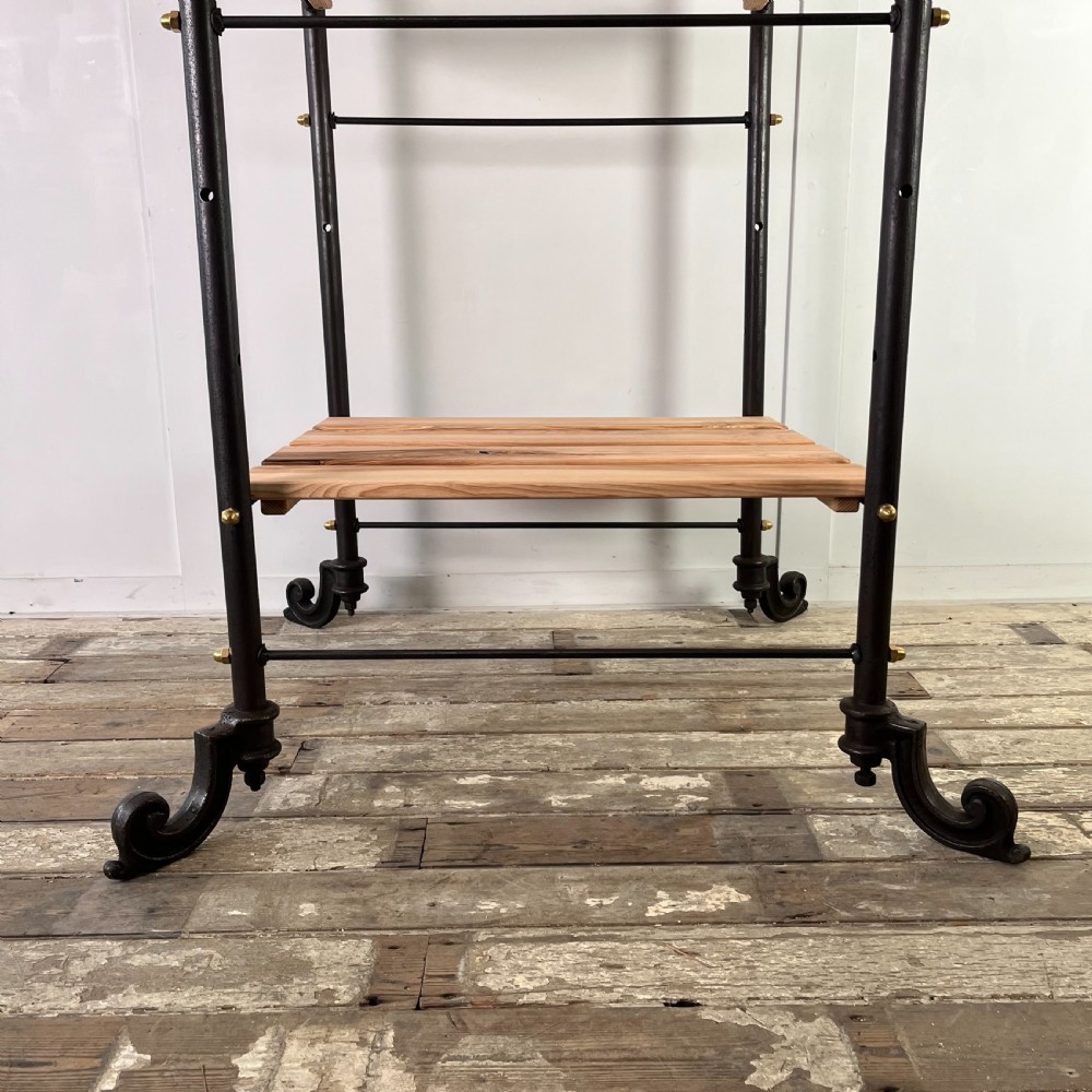 A tall Victorian-style black metal frame display stand featuring three timber slatted shelves, decorative brass finials at the top corners, and scrolled foot supports. View III