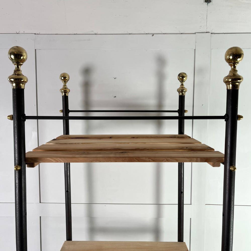 A tall Victorian-style black metal frame display stand featuring three timber slatted shelves, decorative brass finials at the top corners, and scrolled foot supports. View VI