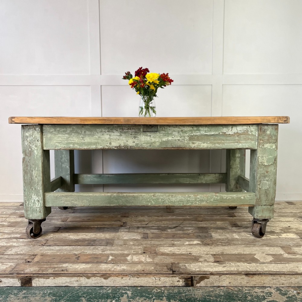 An early 20th-century pine work table featuring a natural scrubbed wood top and a distressed sage green painted frame supported by heavy cast iron castors.
