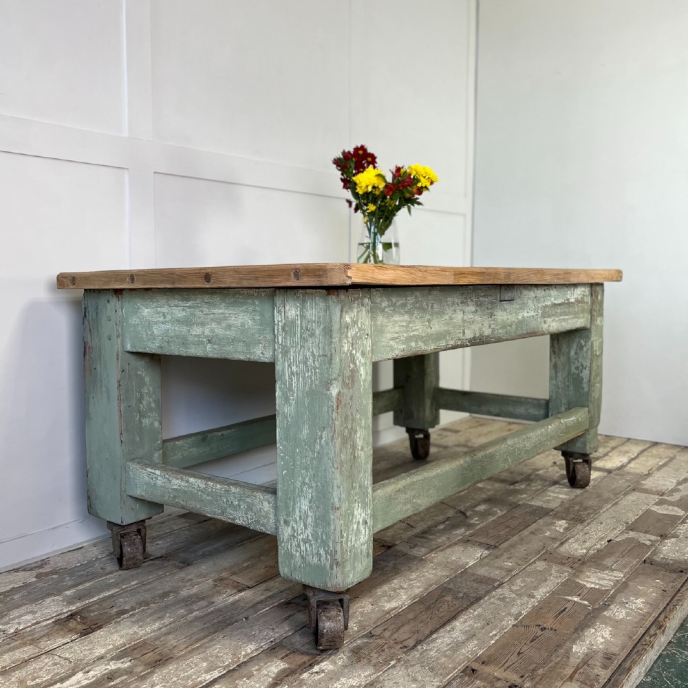 An early 20th-century pine work table featuring a natural scrubbed wood top and a distressed sage green painted frame supported by heavy cast iron castors. View II
