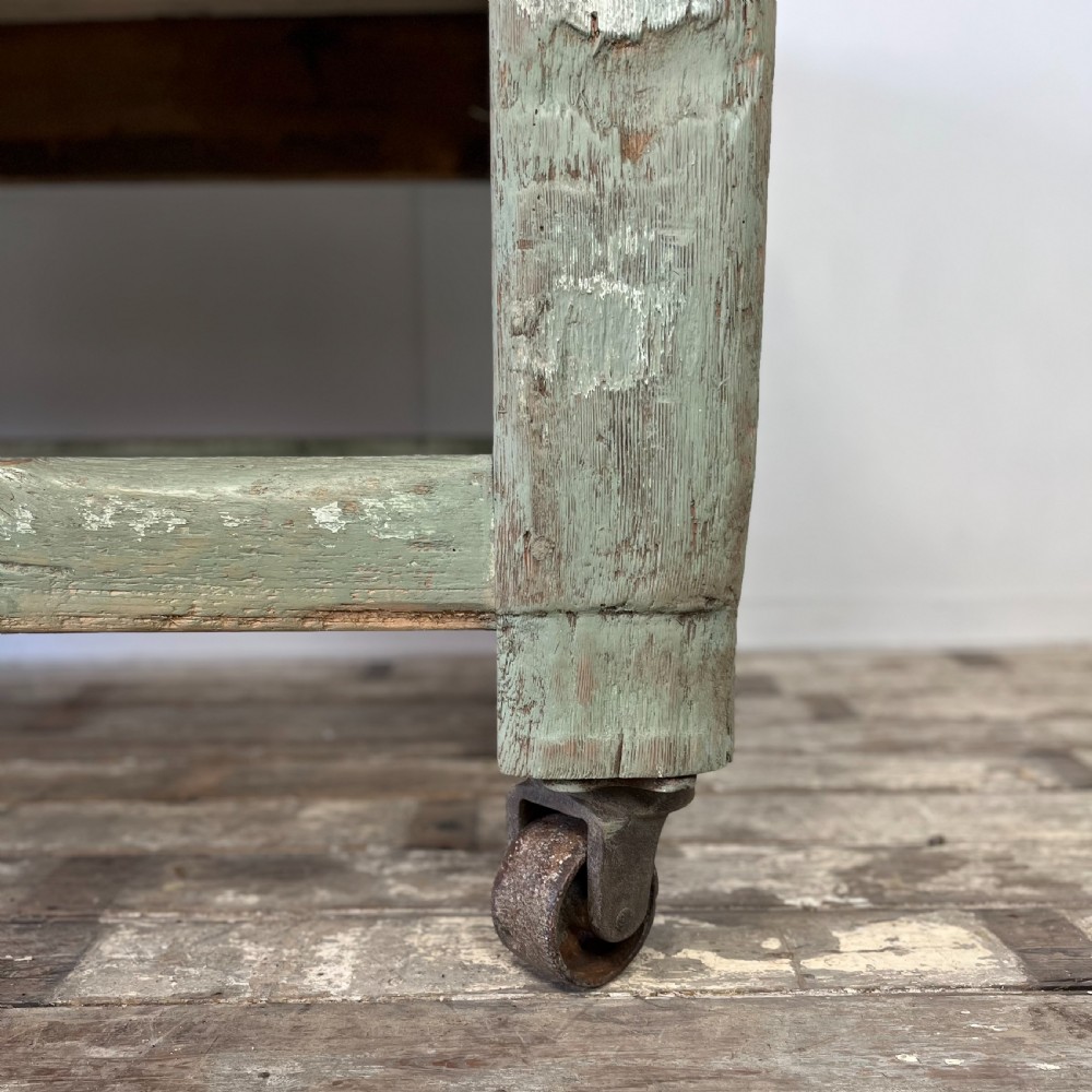 An early 20th-century pine work table featuring a natural scrubbed wood top and a distressed sage green painted frame supported by heavy cast iron castors. View III