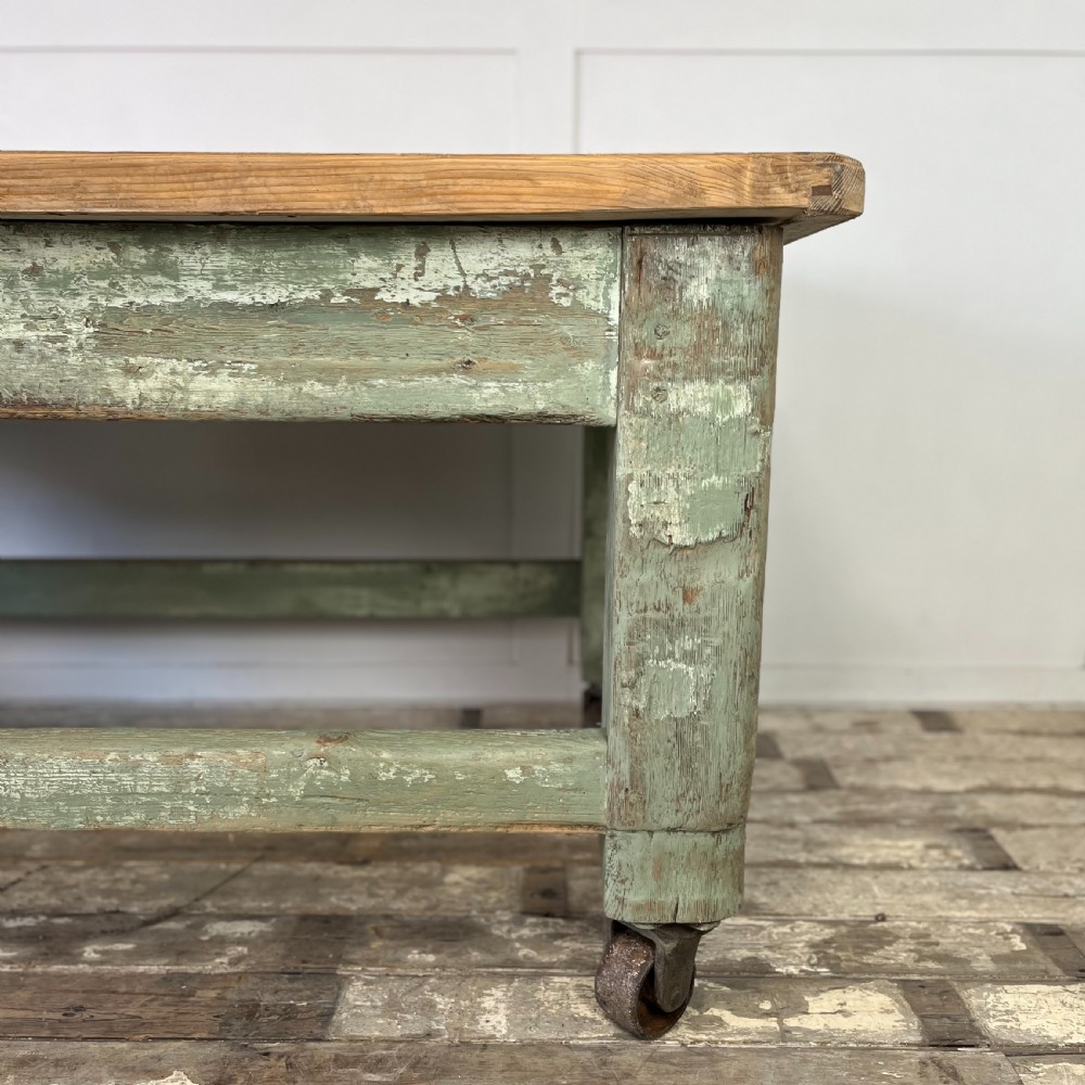 An early 20th-century pine work table featuring a natural scrubbed wood top and a distressed sage green painted frame supported by heavy cast iron castors. View V