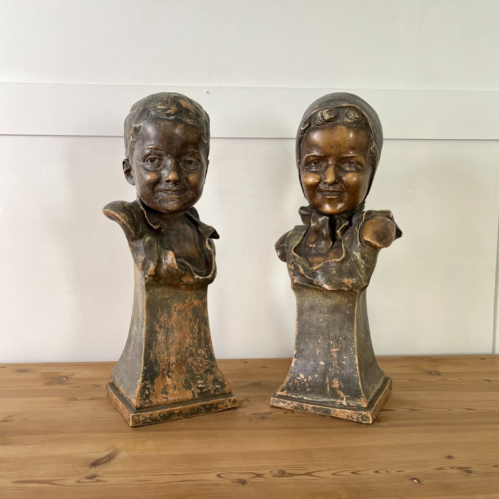 A pair of early 20th-century terracotta busts depicting a young boy and girl with aged bronze-effect finishes on integrated square pedestals.