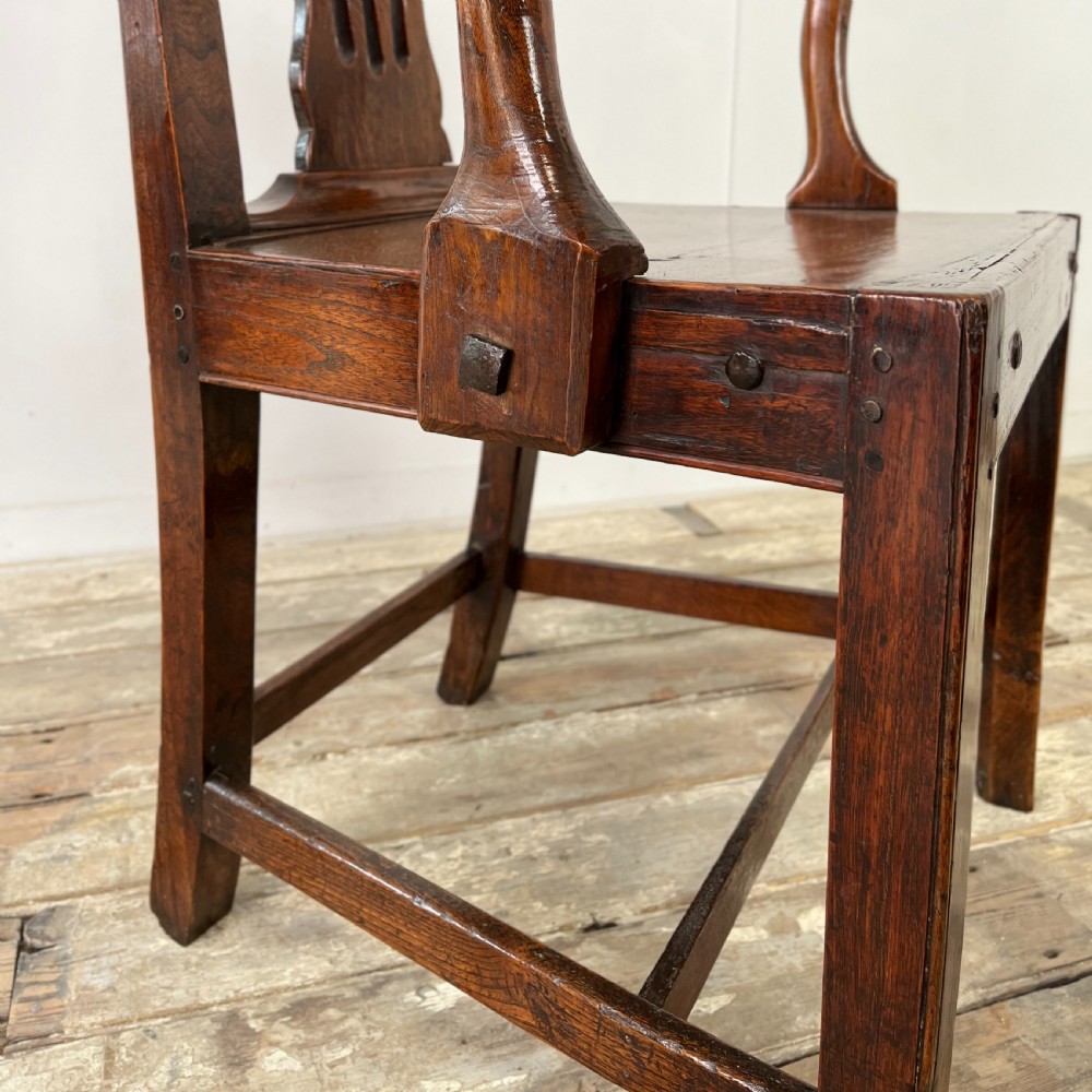 A dark brown early nineteenth-century elm armchair featuring a shaped top rail, a pierced vertical back splat, curved armrests, a solid timber seat, and straight legs joined by stretchers. View XVI