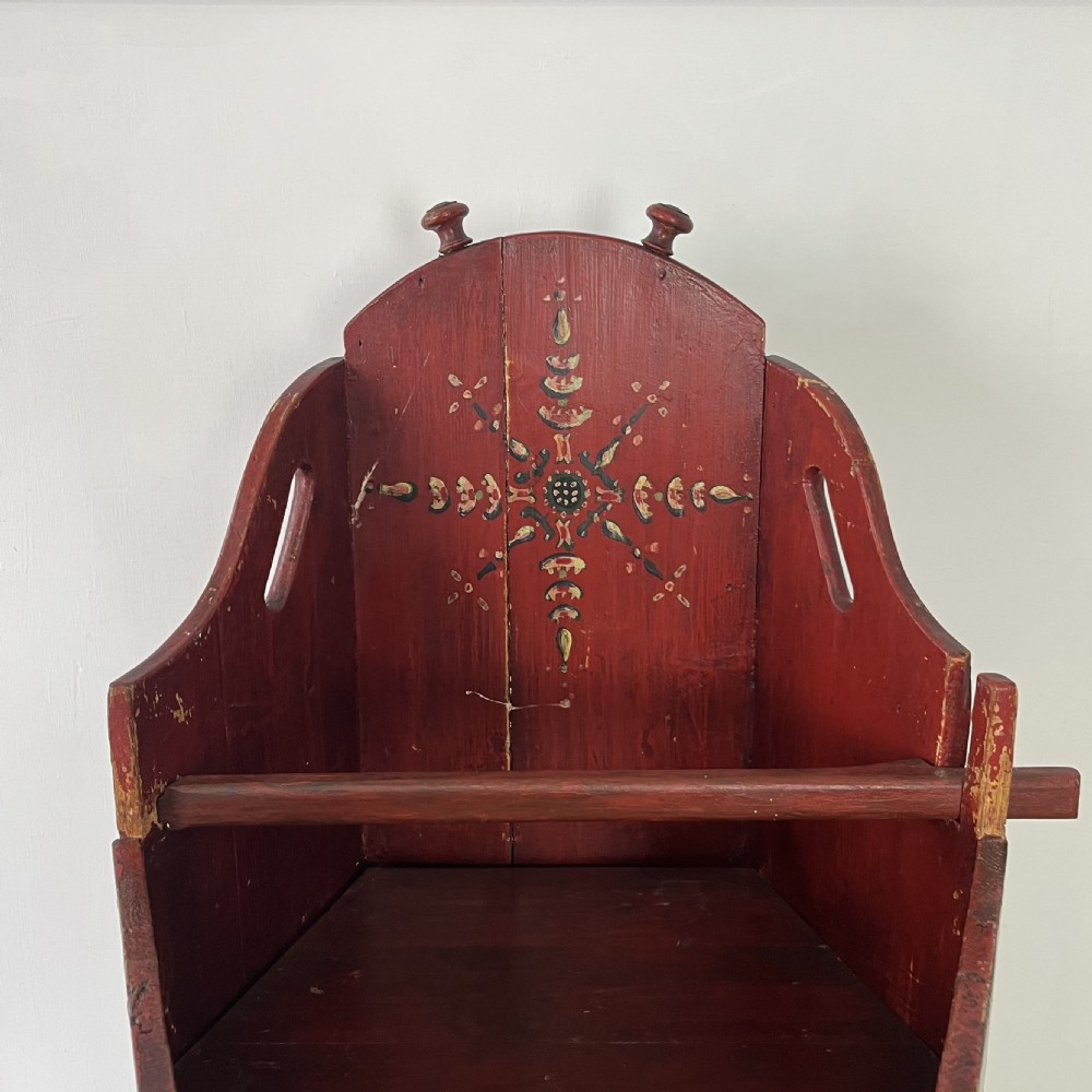 A 19th-century European folk art childs high chair featuring a dark painted enclosed cupboard base and a crimson shaped backrest decorated with hand-painted floral motifs. View V