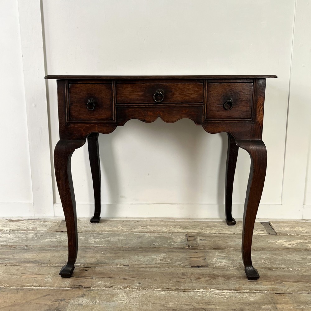 An early 18th-century dark oak lowboy table featuring three drawers, a scalloped apron, and four cabriole legs. View II