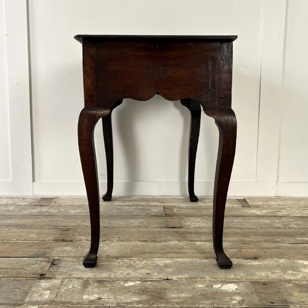 An early 18th-century dark oak lowboy table featuring three drawers, a scalloped apron, and four cabriole legs. View X